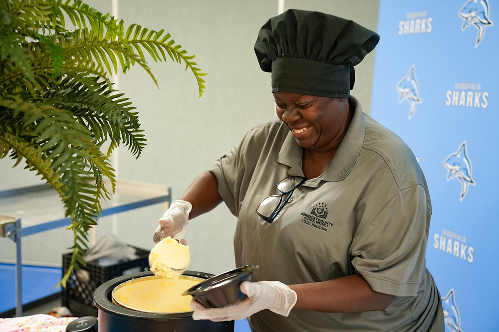 A woman in a gray shirt and chef hat, wearing gloves, mixes food in a bowl.