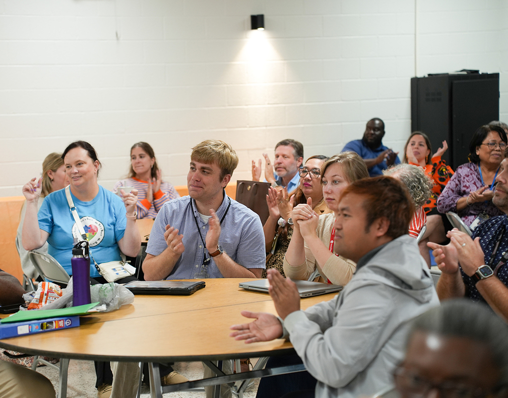A group of people sitting in a circle around a table, clapping. A person stands near a black speaker.