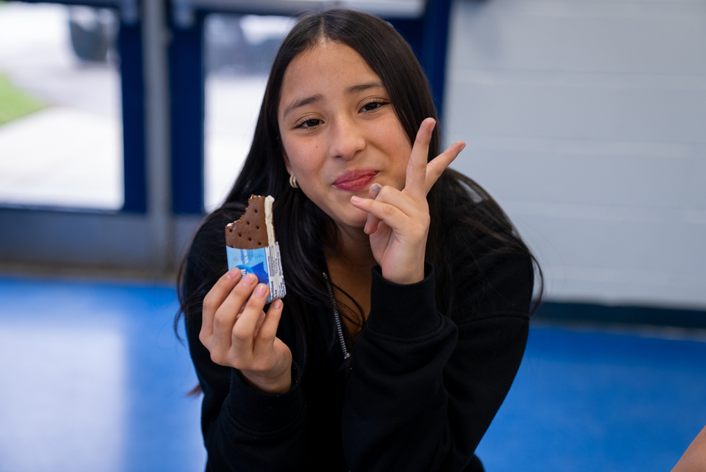 A woman with black hair holds an ice cream sandwich, posing with a peace sign in a room with blue flooring.