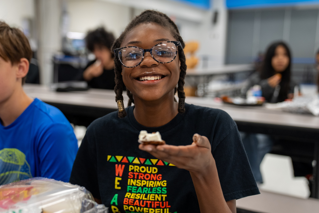Girl in glasses holds a sandwich and smiles at the camera. Other children sit at a table behind her.