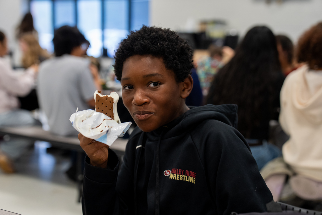 A young boy in a black hoodie sits at a table and holds an ice cream sandwich.