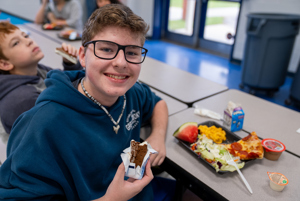 A boy wearing glasses and a hoodie smiles, holding a dessert. Behind him, a boy looks upward. A table has food, utensils, and trash bins.