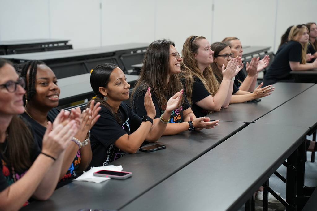 Women sitting at a desk clap while wearing black T-shirts. The desk is long and black.