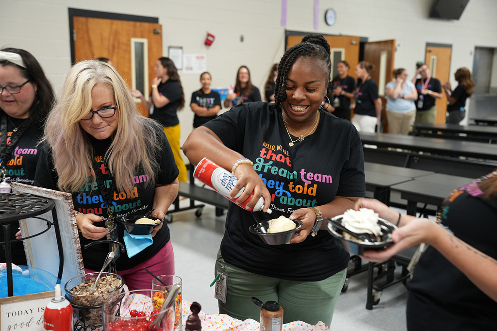 Several women in black t-shirts prepare desserts at a table in a school cafeteria. A clock hangs on the wall.