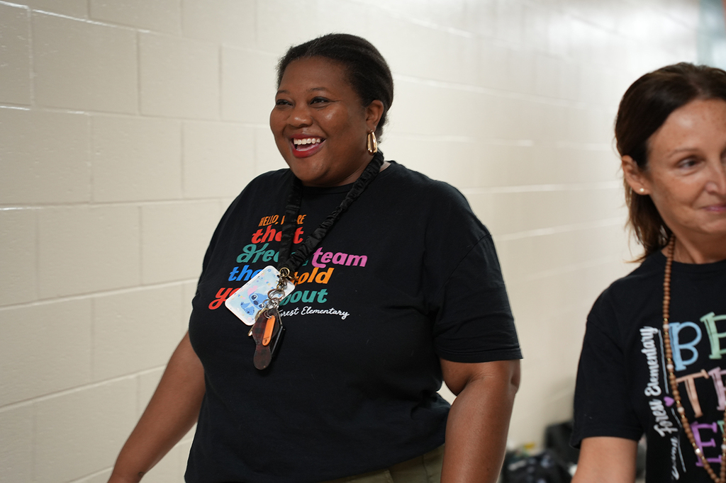Two women in black shirts stand in a hallway. One wears a necklace and ID. Both smile.