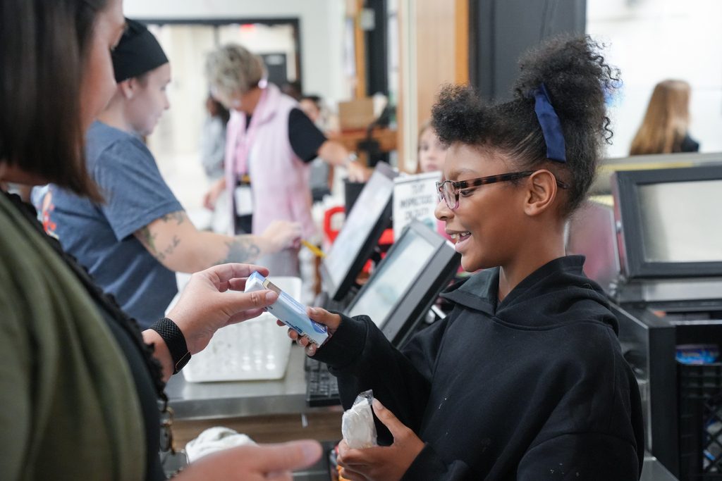 A young girl with glasses holds a cellphone and smiles, interacting with a woman. Behind them, blurred figures stand at a counter.