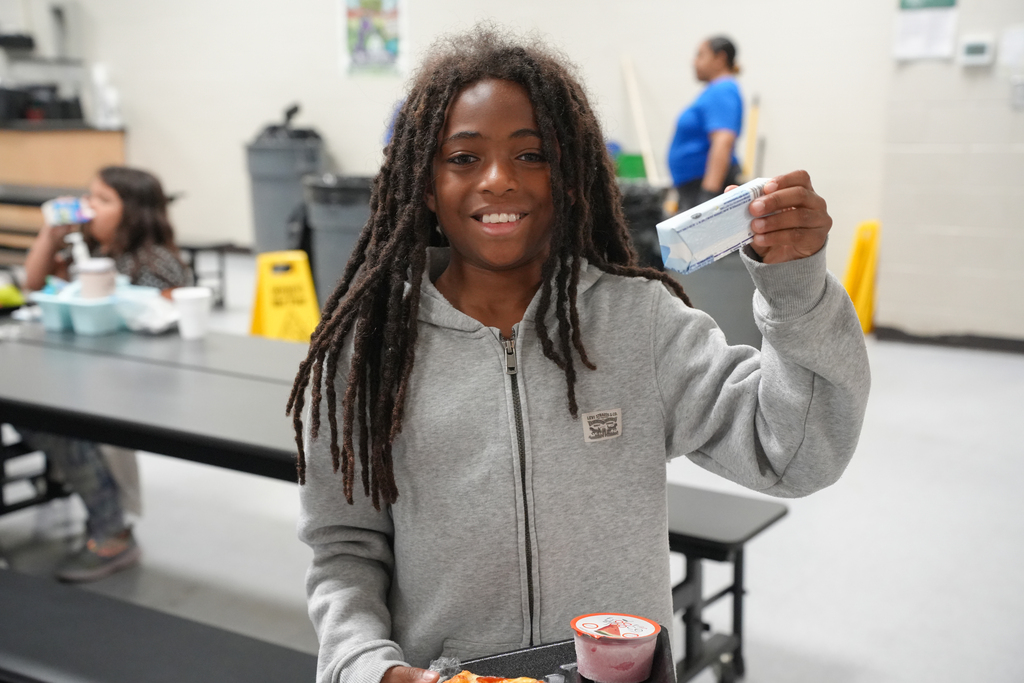 A girl with dreadlocks in a gray hoodie holds up a lunch tray with food in a cafeteria.
