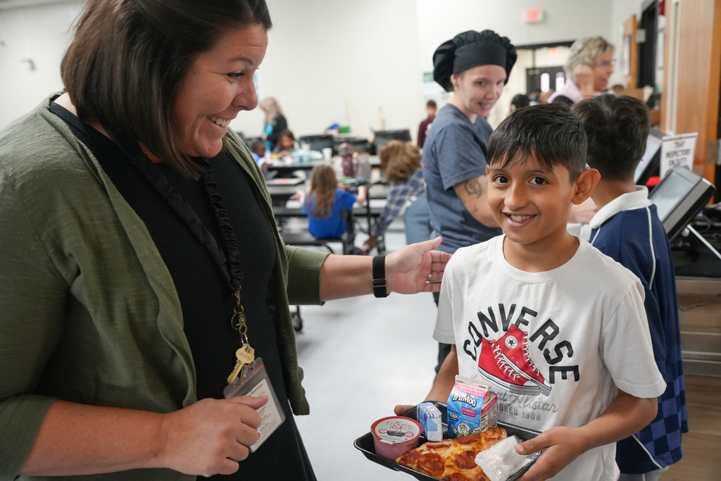 A woman hands a tray with food to a smiling boy in a cafeteria. Other children and adults are in the background.