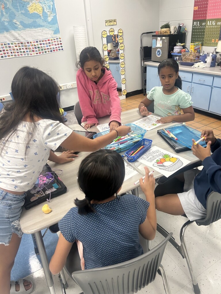 Four children seated at a table with art supplies. One holds a marker. Another is wearing a pink hoodie.