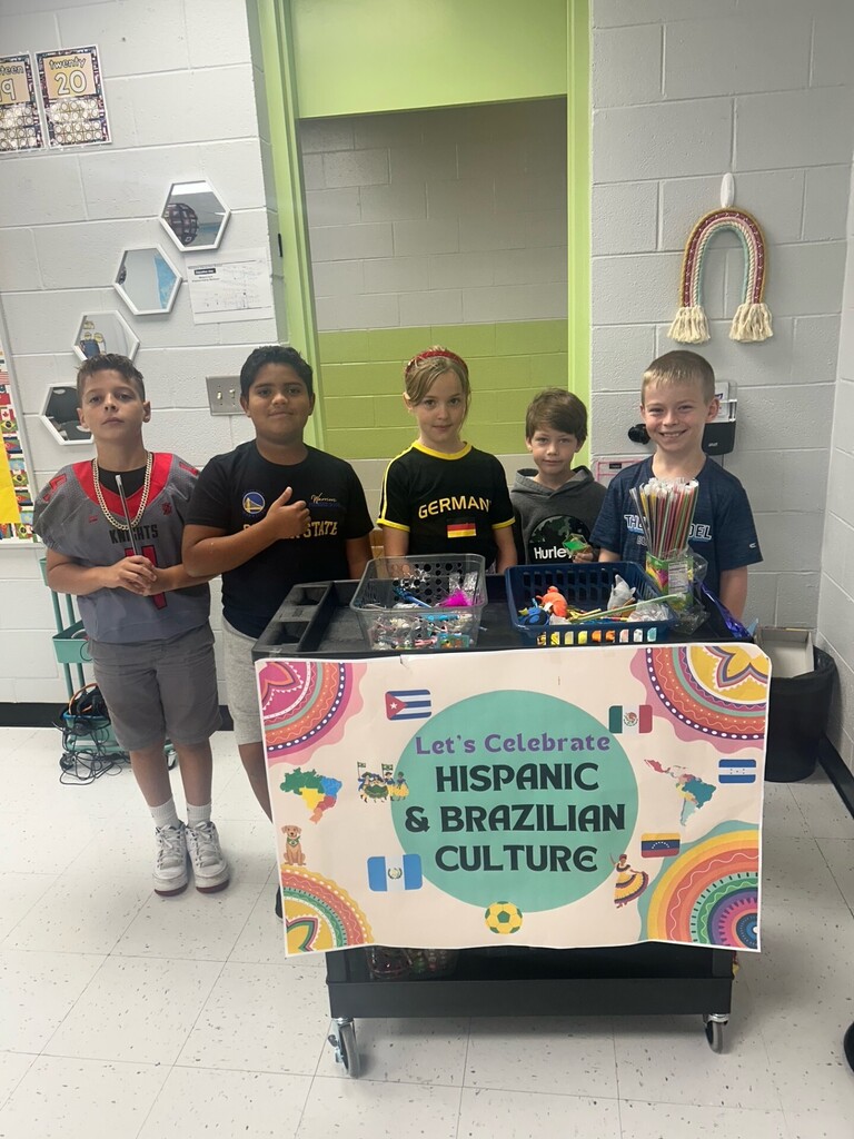 Five children stand behind a table with a sign that says "Let's Celebrate Hispanic & Brazilian Culture." They are in a classroom with white walls and a door.