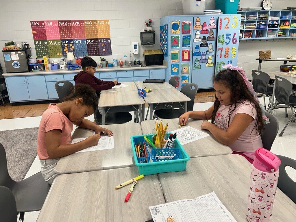 Two children sit at desks, working on papers, in a classroom with blue cabinets and shelves.