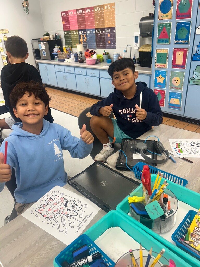 Two children in blue hoodies sit at a table with a laptop and school supplies.