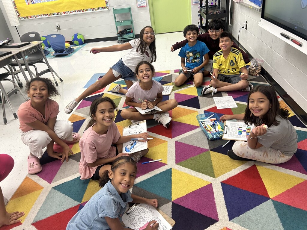 A group of young students sitting on a multicolored carpet, engaged in classroom activities.