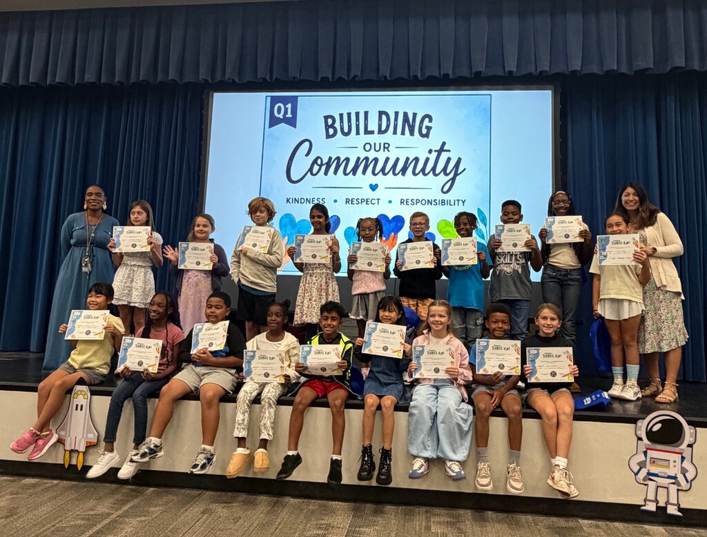 A group of children holding certificates, seated on a stage with a backdrop displaying "BUILDING OUR COMMUNITY."