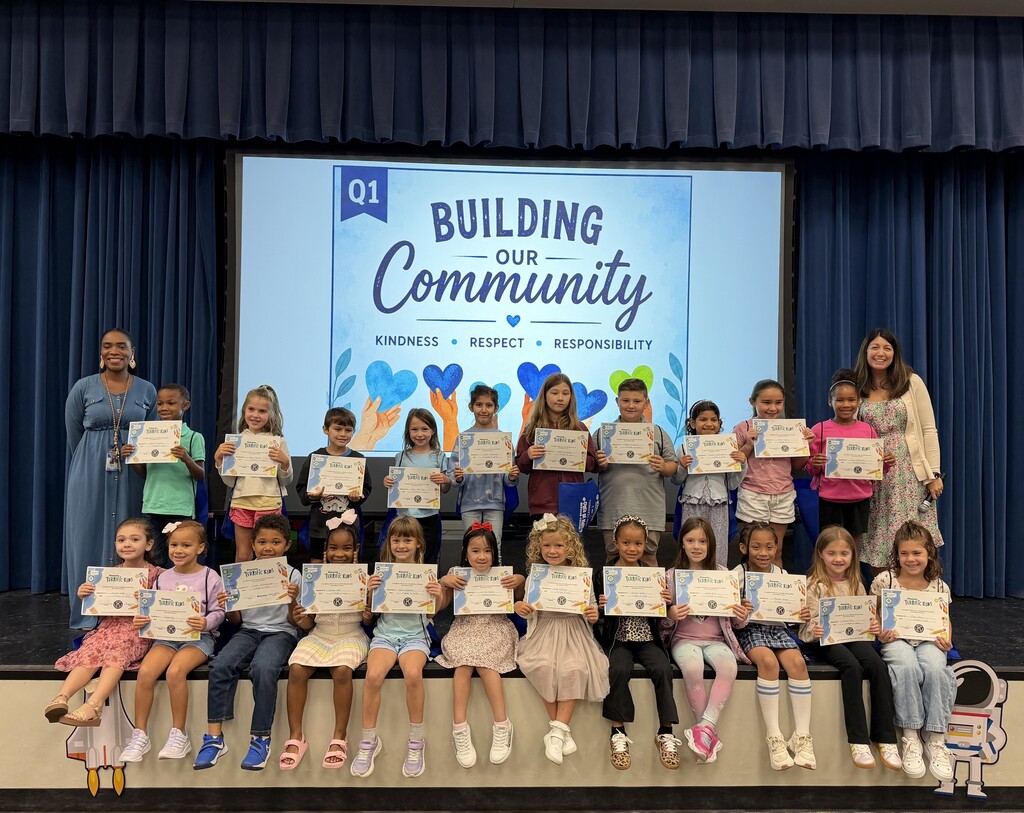 A group of children and two adults holding certificates in a classroom with a large screen.