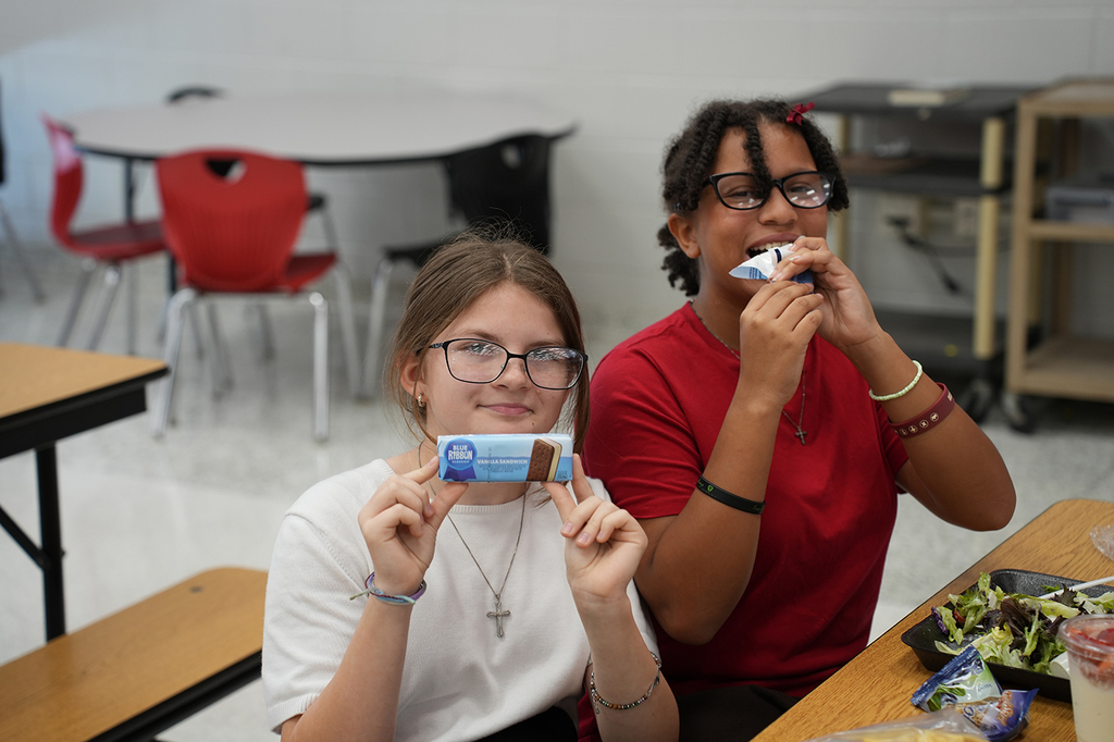Two girls in glasses sitting at a table. One holds a bar and the other eats it.