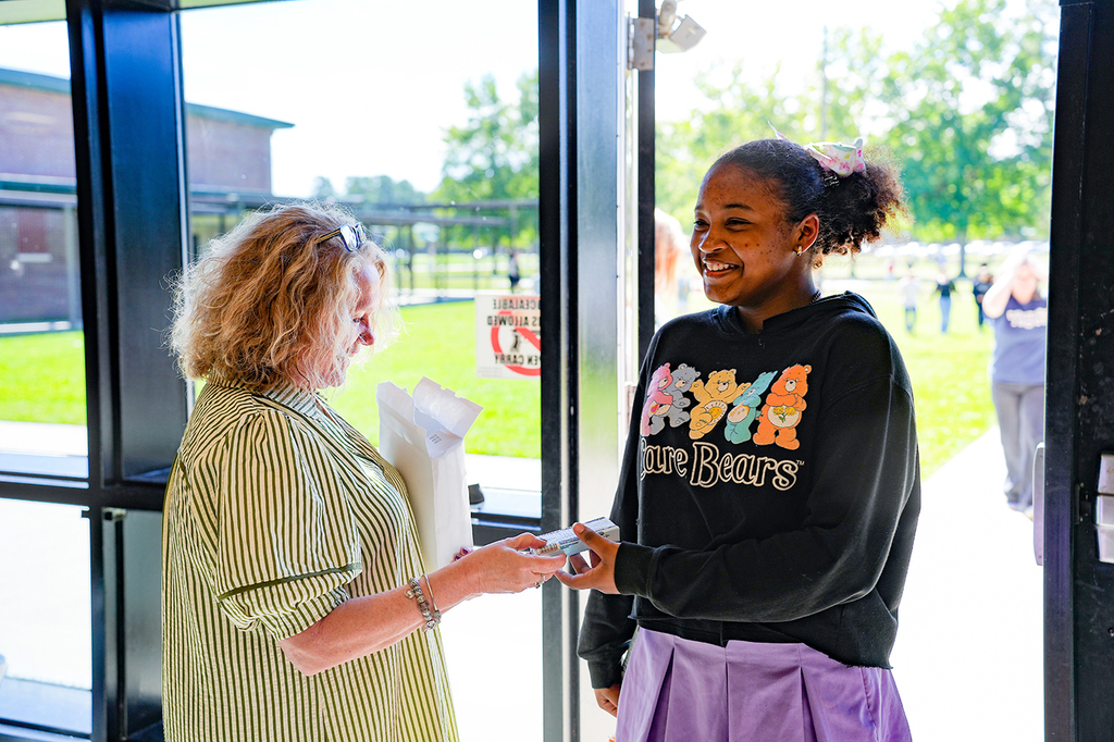 A woman hands a paper to another woman near a glass door with a grassy area and trees in the background.