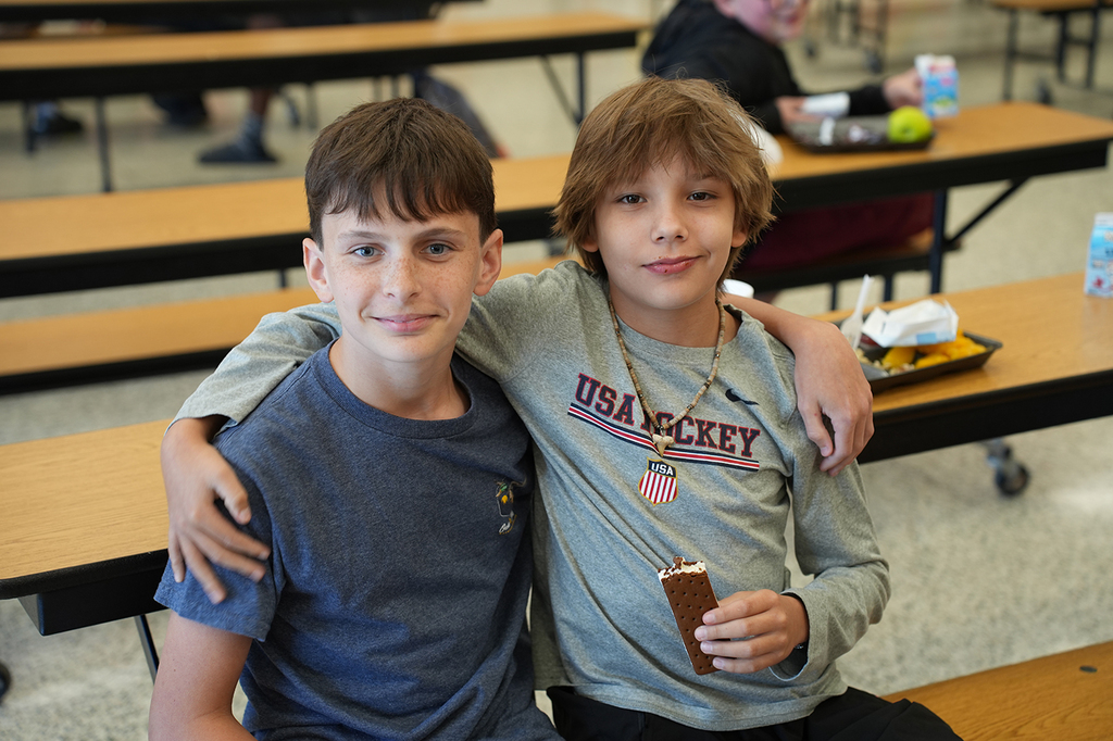 Two boys seated at a cafeteria table, one holding a popsicle. One boy has his arm around the other.