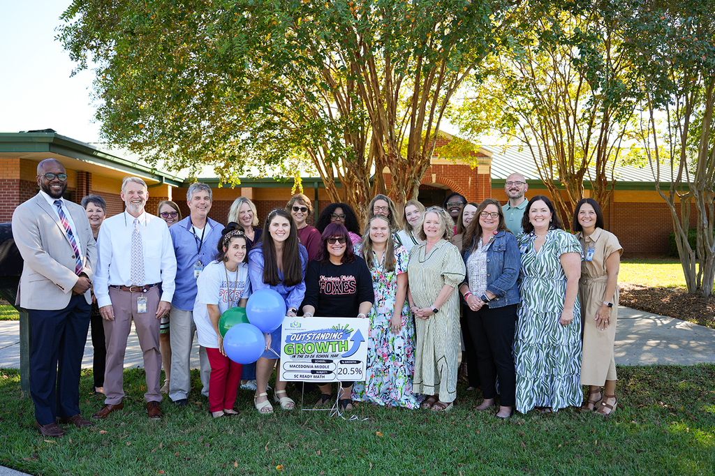 A group of people stand together on grass, holding a banner and balloons. Behind them, there are trees and a building.