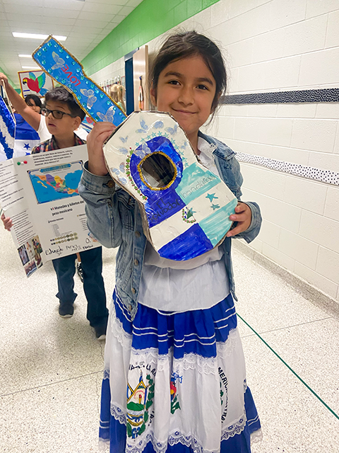 Girl wearing a colorful dress, holding a decorated guitar, in a school hallway.