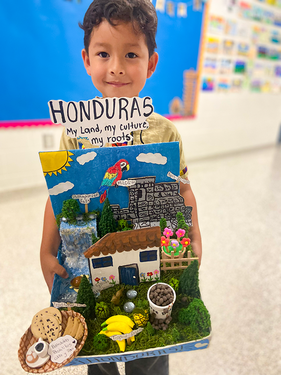 A child holds a box labeled "HONDURAS" with a mini house, waterfall, parrot, and cookies.