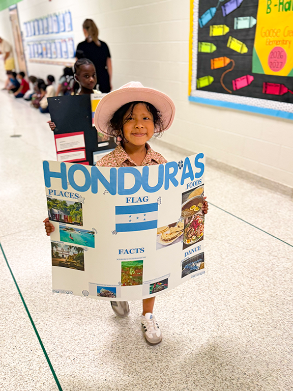 Child wearing a hat holds up a display with information about Honduras. A classroom wall is behind the child.