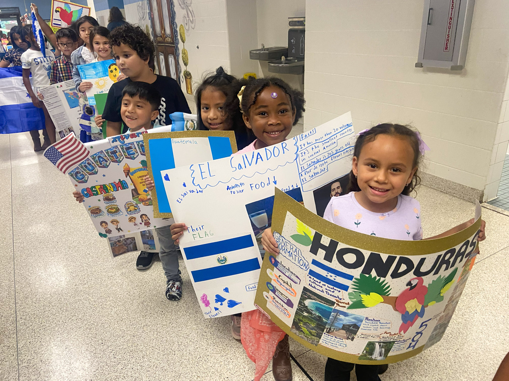 Multiple children holding up signs with countries' names. Background is a hallway with children in the distance.