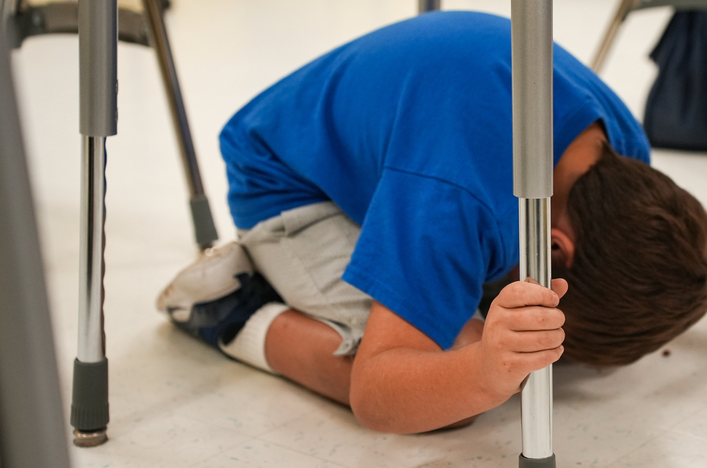 A boy, leaning on a desk, appears distressed with his head on the pole in a classroom.