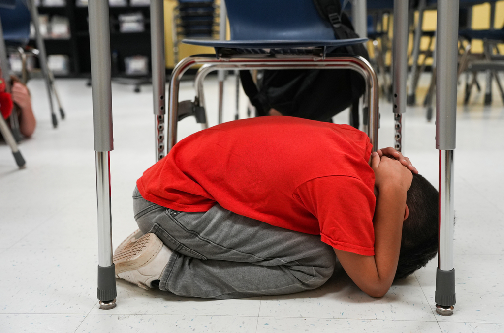 A child sits on the floor with head down and hands covering face in a classroom.