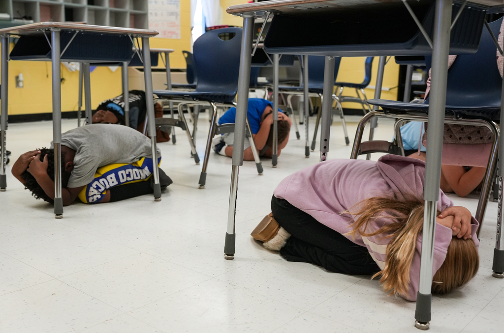 Several students are on the floor, heads down, in a classroom. Yellow and white walls. Chairs are arranged in rows.