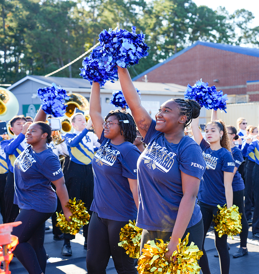 Cheerleaders wearing blue shirts hold blue and gold pom-poms in an outdoor setting with a school building and trees.