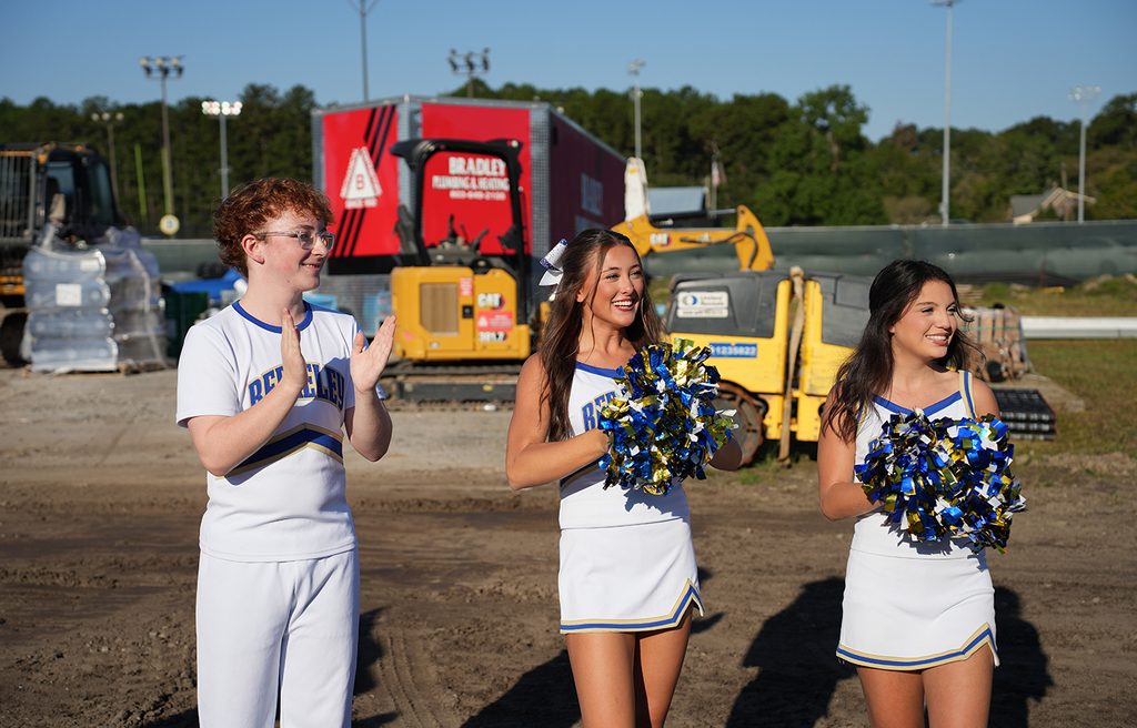 Three cheerleaders in white uniforms stand on a dirt field with machinery in the background.