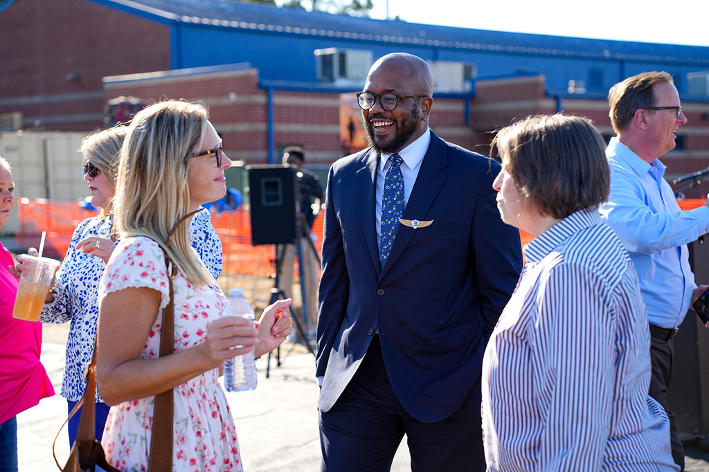 A man and three women in business attire converse. Behind them, a building and a tripod stand.