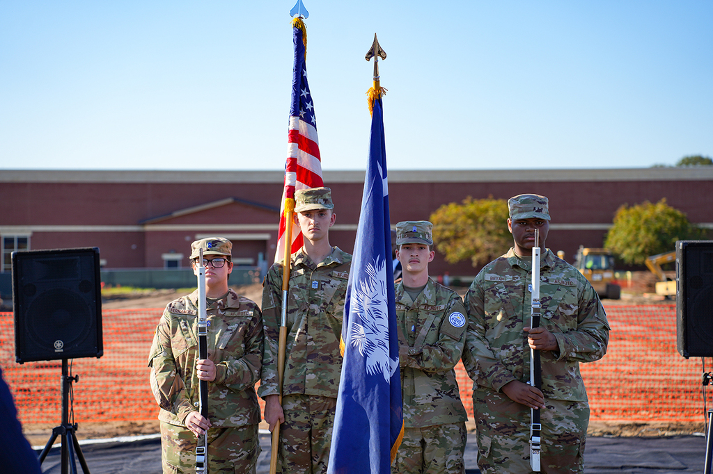 Four soldiers in military uniforms holding flags, standing in front of a construction site with a building and trees in the background.