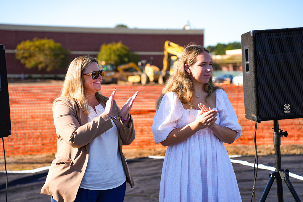 Two women clapping hands. A construction site with machinery visible in the background. An orange barrier surrounds the area.