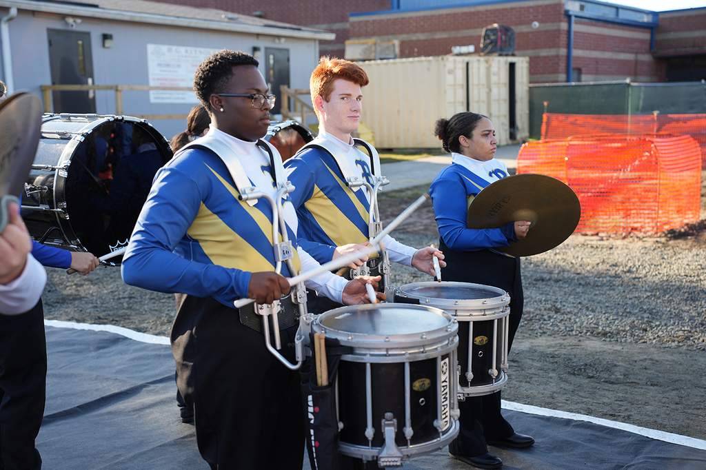 A group of individuals in blue and yellow uniforms play drums outdoors. Behind them, there are buildings and orange fencing.