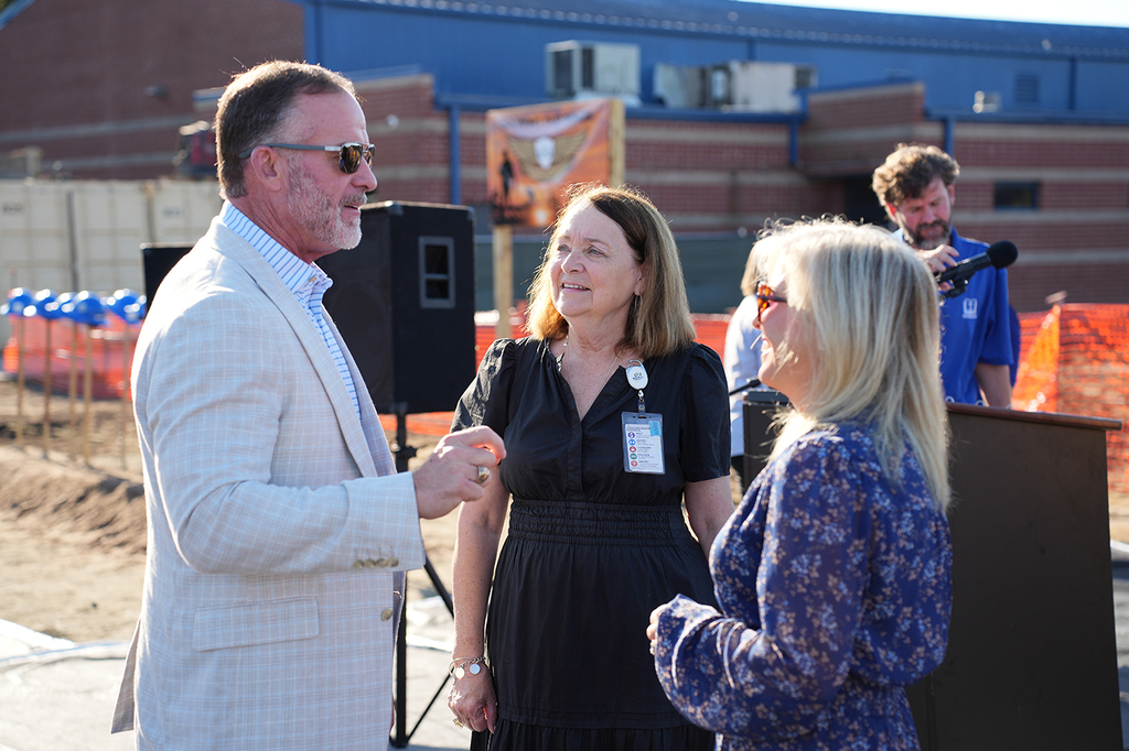 Three adults converse near a podium. One man and two women. Behind them, a building, fence, and construction site.
