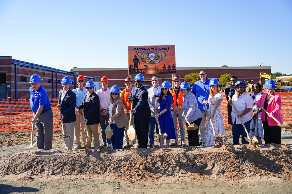 People in hard hats gather at a construction site. A banner is displayed in the background.