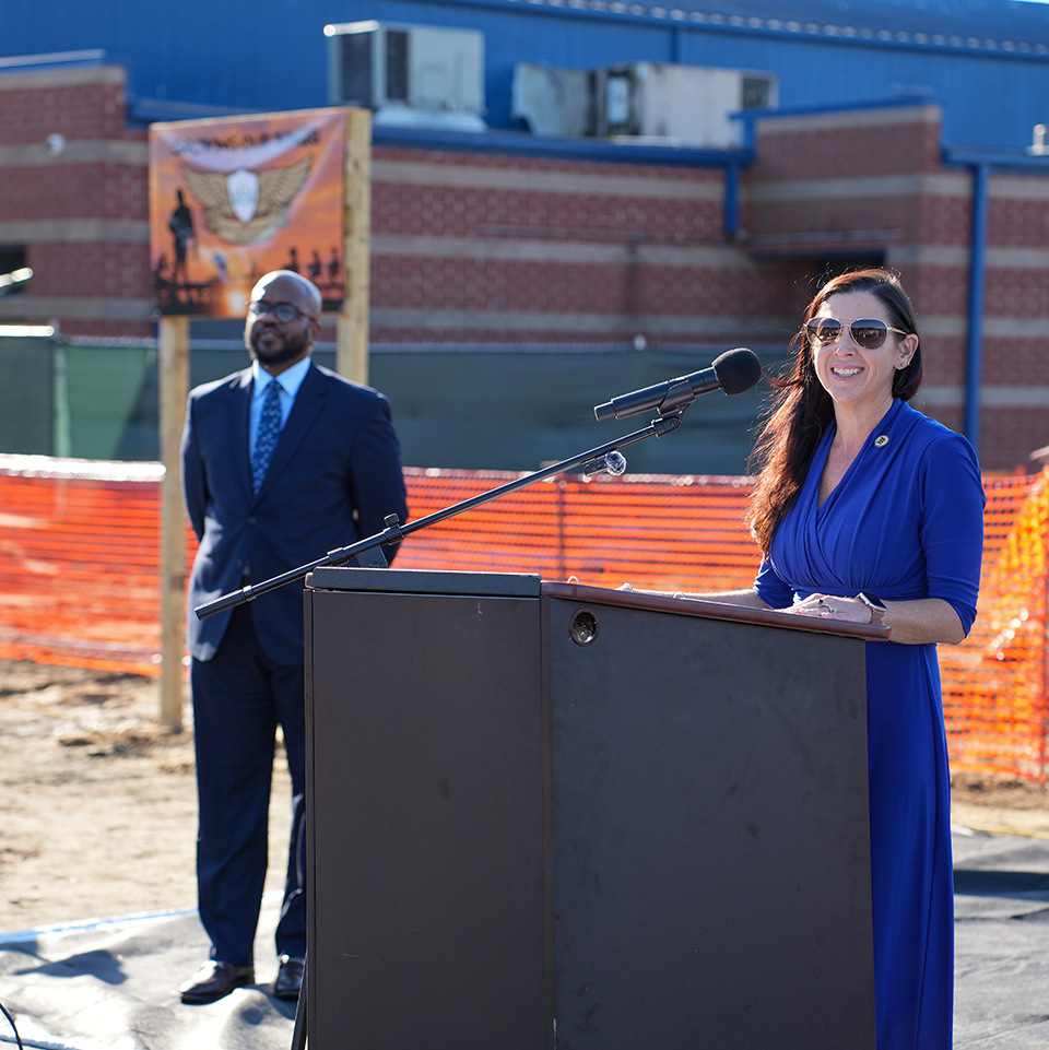 A woman in a blue dress stands at a podium with a microphone, while a man in a suit stands behind her. Construction barriers and a building are in the background.
