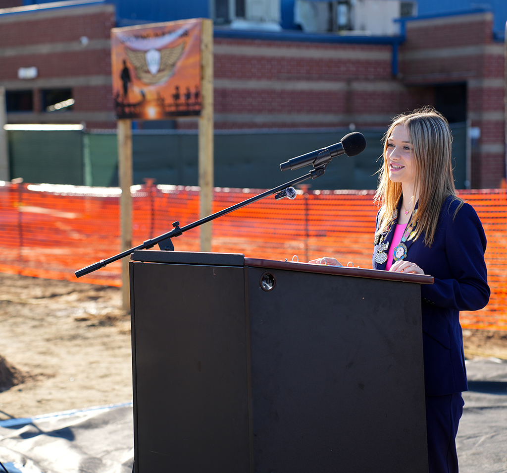 Woman with long blonde hair stands at podium with microphone, in front of a construction site.