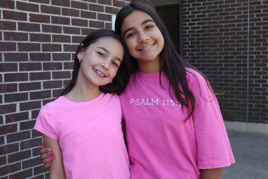 Two young girls smile and pose for a photo. Both wear matching pink shirts with white text.