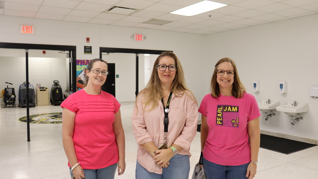 Three women in pink shirts and glasses stand in a white hallway. A sink with two faucets is on the wall.