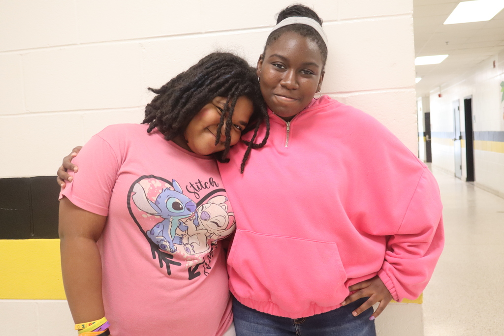Two young girls stand together, one wearing a pink shirt with a cartoon character, the other in a pink sweatshirt. They are inside a school hallway.