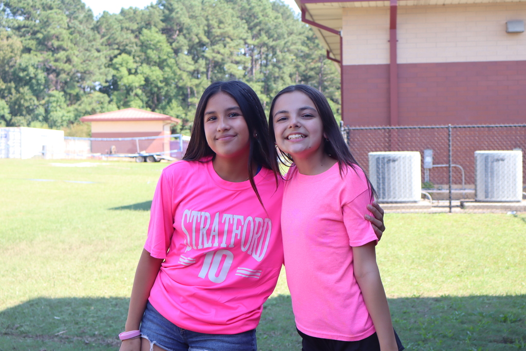 Two girls wearing pink shirts stand together in a grassy area, smiling. Behind them is a building with air conditioners.