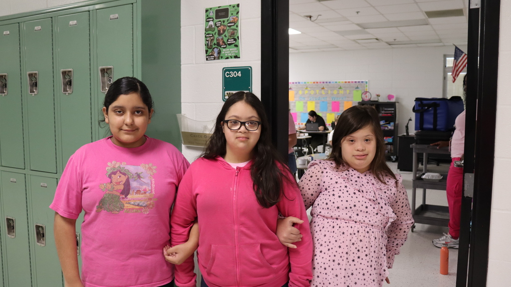 Three girls in pink shirts or pajamas standing in front of lockers, with a classroom visible through the open door.