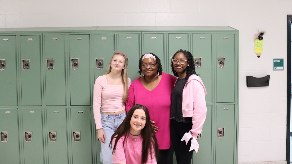 Four people in pink shirts standing in front of a row of lockers with a green door.