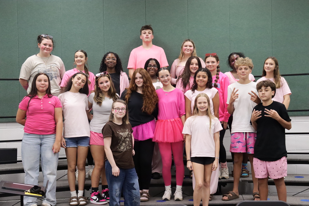 Group of students standing on stage wearing pink and white shirts, shorts, and skirts.