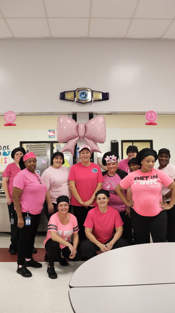 A group of people in pink shirts and black pants standing together, smiling, with a pink balloon above.