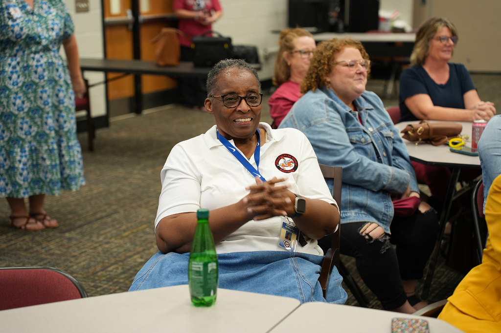 Five women seated at tables in a classroom, one woman with a lanyard smiles and gestures with hands.