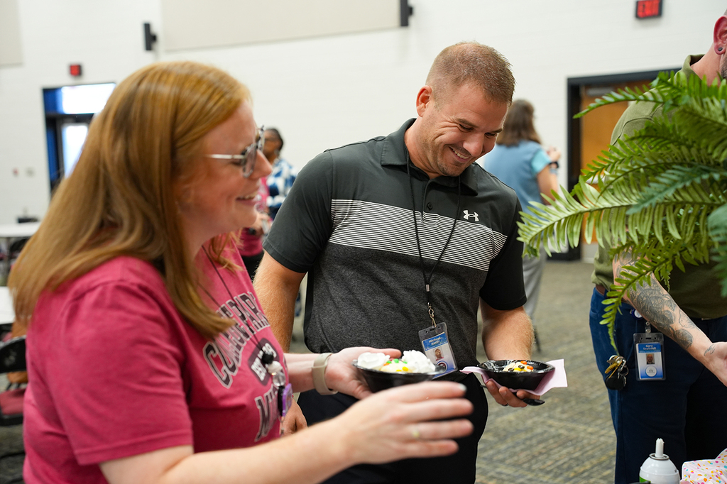 Two adults, one smiling, exchanging bowls of food in a large room. One holds a nametag.